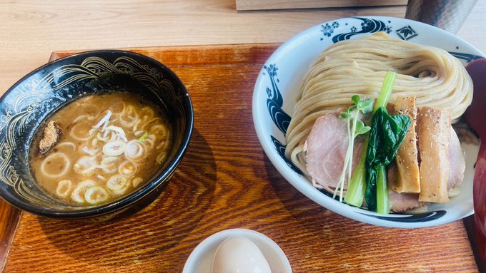 The image shows a wooden table with two bowls of food, one containing noodles and the other soup. The bowl of soup has a white egg floating in it. There are also some vegetables present in both bowls. A small plate can be seen on the side as well. The background is blurred out, but there appears to be a window or another source of natural light that illuminates the scene.