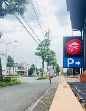 The image shows a person running down a street, wearing a white shirt with the Pizza Hut logo on it. The person appears to be enjoying their run while passing by a pizza shop called Pizza Hut. In the background, there are several trees lining the street, adding a touch of nature to the urban setting.