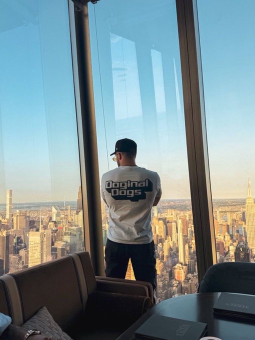 A man wearing a white 'Doginal Dogs' t-shirt and dark cap stands at floor-to-ceiling windows overlooking the New York City skyline, with the Empire State Building visible.