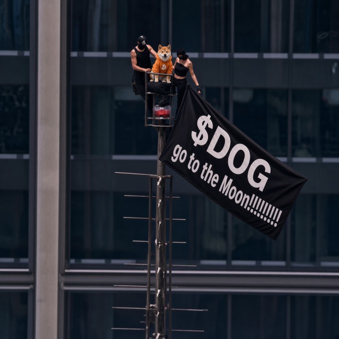 Two people stand on a high platform with a Shiba Inu mascot in an orange “6” shirt, while a black flag reads “$DOG go to the moon!!!!!!!!” in a playful reference to the post’s call for the $DOG ticker to surge.