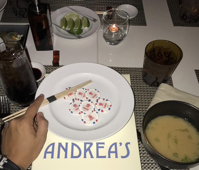 A hand holds chopsticks over a white plate featuring sushi-shaped chips with "5000" printed on them, placed on a placemat reading "ANDREA'S," accompanied by a bowl of miso soup and a candle-lit table setting. The image relates to the "Gn bros" caption by showcasing an intimate, food-centered dinner for two.