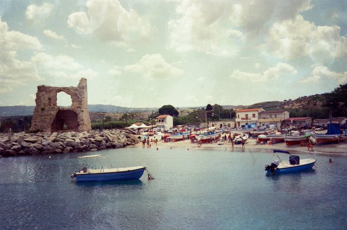 A scenic harbor view featuring two small boats floating on calm water in the foreground, with an ancient stone tower ruin on the rocky shore and a coastal village with beachgoers in the background under a cloudy sky.