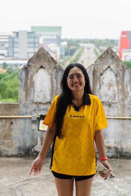 A smiling woman with long dark hair and glasses stands outdoors holding a Nintendo Switch, wearing a yellow 'BINANCE' branded polo shirt, with city buildings visible in the background.