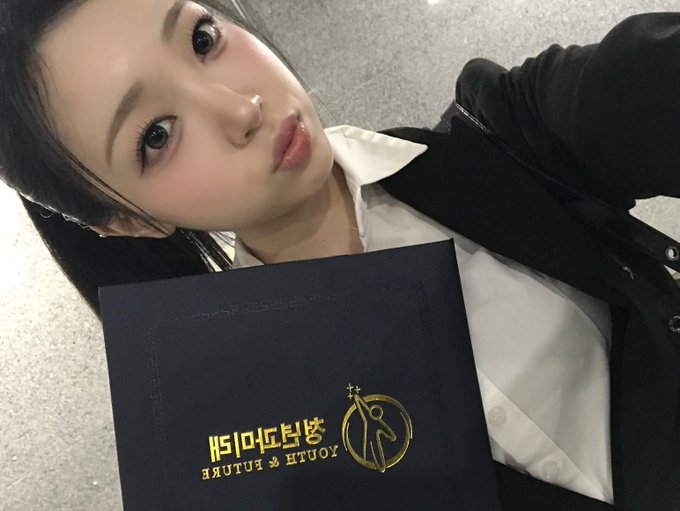 A selfie of a young woman holding a black folder with the 'YOUTH & FUTURE' golden logo, wearing a white collared shirt under a black jacket.