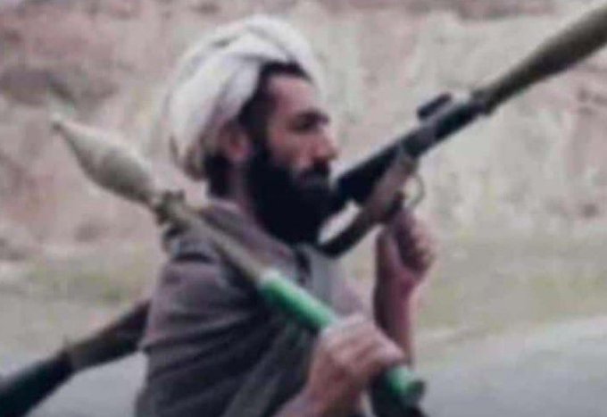 A blurry image of a bearded man wearing a white head covering, carrying what appears to be a long weapon or rifle across his shoulders in an outdoor rocky setting.