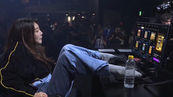 A young woman relaxes with her feet up at a computer monitor displaying multiple sneaker product pages, likely bidding in real-time. The setup, complete with a water bottle and attentive crowd in the background, humorously illustrates the dedication—and casual comfort—of a serious sneaker collector.