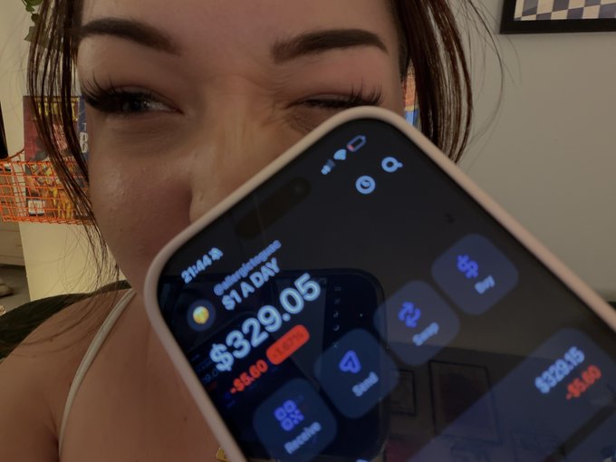 A close-up selfie shows a woman holding a smartphone displaying a stock portfolio app with a Bitcoin balance of '$329.05' and a daily loss of '-$5.00'.