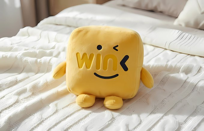 A yellow plush toy featuring the word "win" with a winking smiley face sits on a white knitted bedspread. The cheerful "win" character playfully symbolizes the successful achievement of verifiable carbon storage goals in TRON-automated peatland restoration grants.