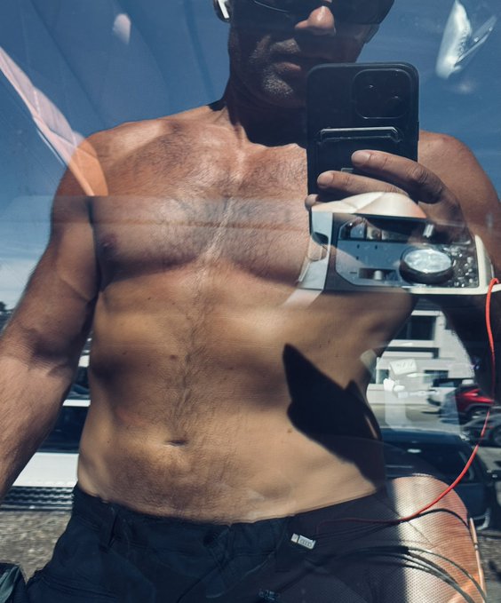 A shirtless, muscular man wearing sunglasses takes a mirror selfie with a smartphone while holding a silver vintage-style camera with a red cable.