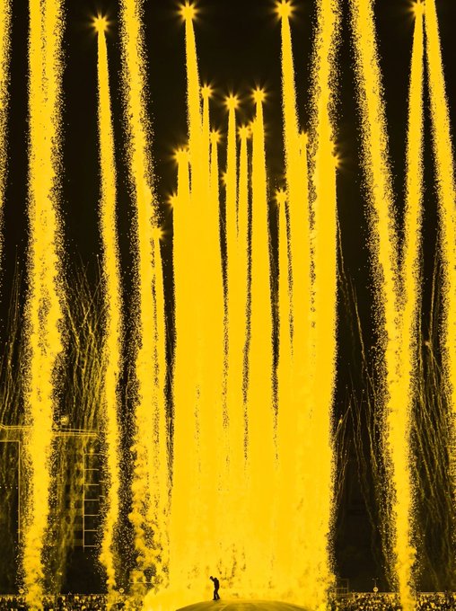 Bright yellow fireworks or pyrotechnics stream downward against a black night sky, with a small figure visible at the bottom.