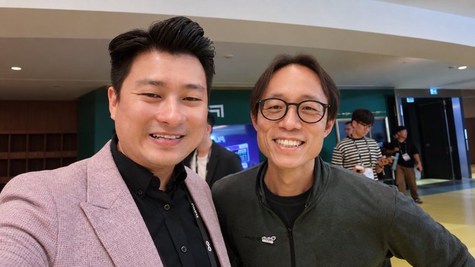 A selfie of two smiling Asian men at an indoor event; the man on the left wears a pink blazer over a black shirt, while the man on the right wears glasses and a dark gray zip-up jacket.