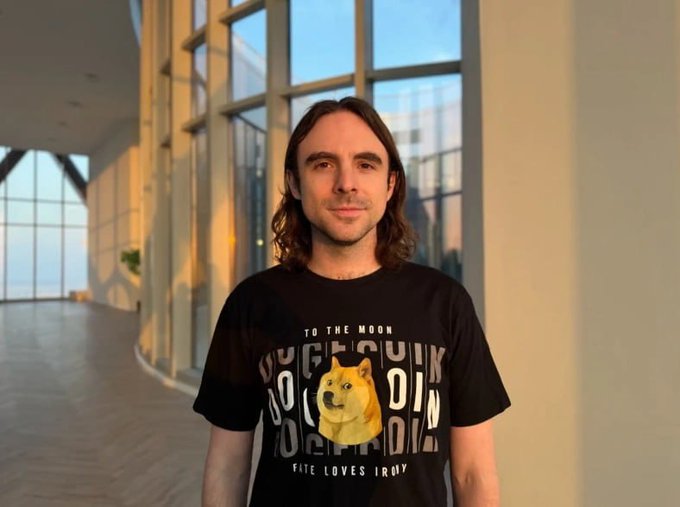 The image features a man standing next to a window with his arms crossed. He is wearing a black t-shirt that has the word "To the moon" printed on it, possibly indicating an interest in space exploration or other moon-related activities. The photo also includes text about a Dogecoin meme and its association with the Dogecoin community.