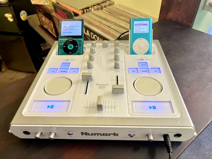 A Numark DJ mixing console sits on a wooden surface with two iPods docked in its top cradle, displaying track information on their screens.