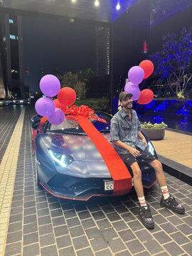 A photograph of a young man sitting on the hood of a Lamborghini sports car decorated with a large red ribbon and balloons.
