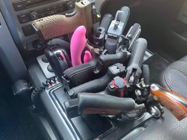 An absurd photo showing a vehicle's center console overflowing with an excessive collection of handguns, pistols, and weapons stuffed into the cup holders and gear shift area.