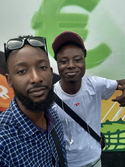 A selfie of two men standing together, one wearing a blue plaid shirt with sunglasses on his head and the other wearing a grey t-shirt and a maroon baseball cap, standing in front of a green mural.