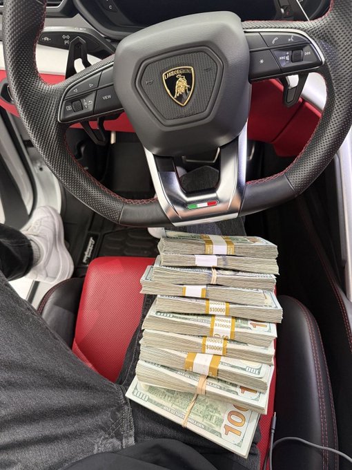 A photo taken inside a Lamborghini showing a person sitting in the driver's seat with their lap covered in banded stacks of US $100 bills, with the steering wheel's Lamborghini logo visible.