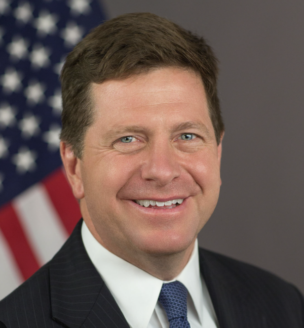 An official portrait of a smiling man in a dark suit with an American flag in the background.