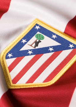 A triangular patch featuring red and white stripes with blue sections adorned by white stars displays a figure with a soccer ball for a head, designed in a style resembling a classic superhero emblem. The image ties into a satirical comment about refereeing decisions favoring Real Madrid, with the logo serving as a playful emblem for Atletico Madrid's perspective on the issue.