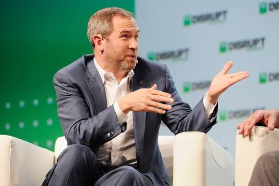 A man in a gray suit gestures while speaking at a Digirati-branded event panel, appearing to discuss topics such as Ripple's recent full MiCA license approval from Luxembourg for operating across EEA markets.
