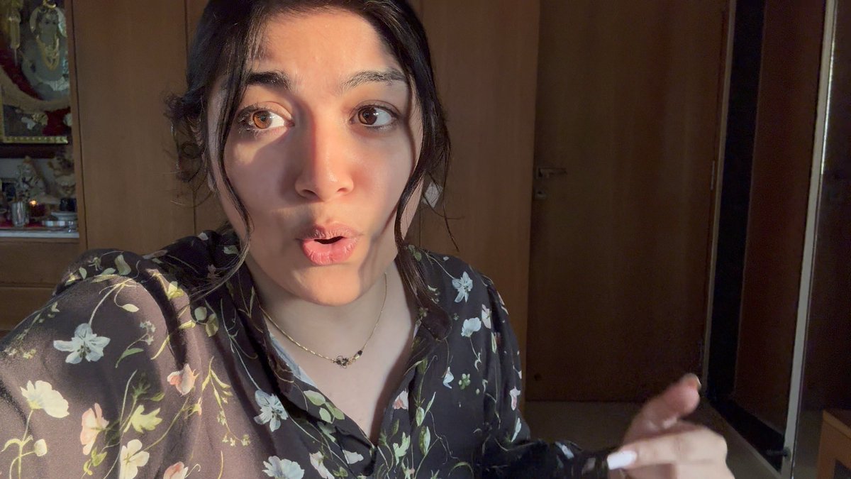 The image shows a young woman with dark hair wearing a floral-patterned blouse and a delicate necklace, speaking directly to the camera in what appears to be an indoor setting with warm wooden doors in the background. Her expressive mid-speech face and casual setting suggest she's recording a personal video message, likely one of the "meet you guys in Bombay" announcements referenced in the post about an upcoming Bitcoin-focused event in India.