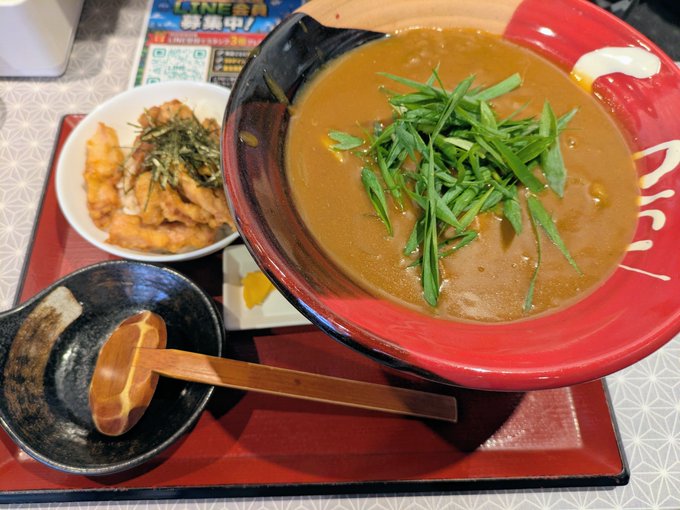 A Japanese meal set featuring a large bowl of thick curry topped with fresh green onions, a smaller bowl of fried chicken or tempura with herbs, and a wooden spoon resting in a black dish, served on a red tray.