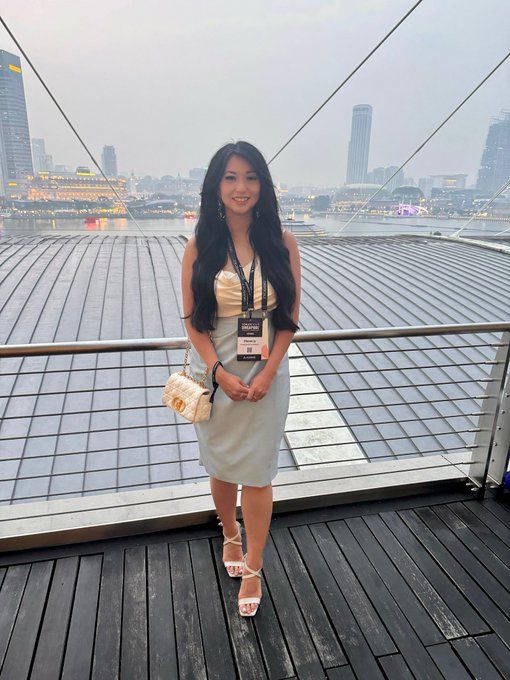 A woman in a light blue dress with a TOKEN2049 lanyard poses on a wooden deck overlooking the Singapore skyline and Marina Bay Sands, capturing the elegant end to Day 1 of the conference.