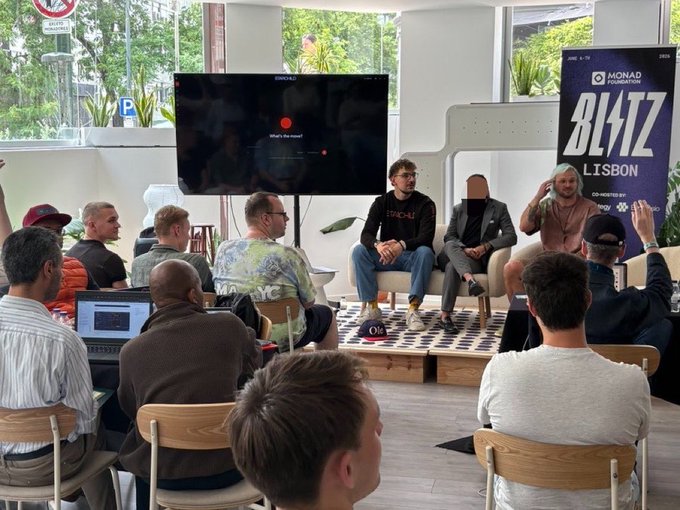 The image shows a group of people sitting around a table, engaged in conversation. There are at least nine individuals present, with one person wearing a baseball cap. The room has a large window that allows natural light into the space, creating an inviting atmosphere for the gathering. A TV screen is visible on the wall behind the group, and there's also a laptop placed nearby.

The people in the image appear to be discussing various topics or sharing personal stories with each other. One person seems particularly interested in what others are saying, as they lean forward and listen attentively. The overall scene conveys a sense of camaraderie and connection among the group members.