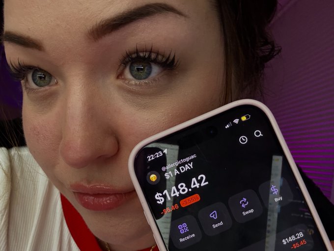 The image features a woman showing off her new cell phone, which has a finance app open that displays a balance of $1.48 for a specific day. The woman is standing next to the display screen with the cell phone in front of her. There are no other people visible within the frame or any text accompanying this scene.