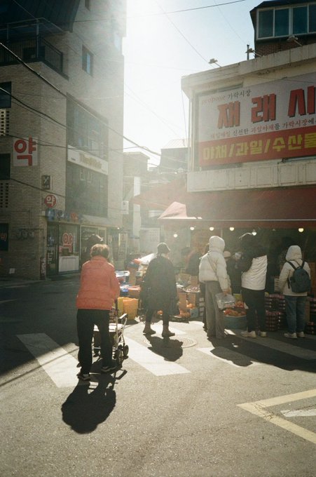 A sunny Korean street scene with people walking past a store with a red sign reading "재래시" (Traditional Market) and advertisements for agricultural products.