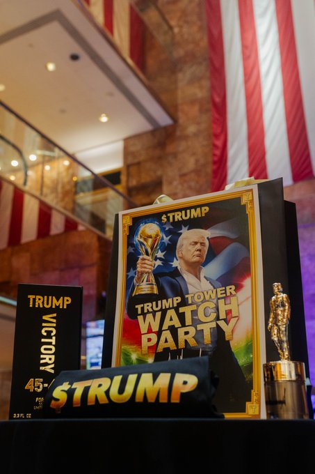 A $TRUMP Trump Tower Watch Party sign featuring a portrait of Donald Trump holding a trophy stands alongside Trump Victory 45-themed merchandise and a golden statuette at an upscale venue. The display connects to the $TRUMP Coin Club World Cup weekend events held across New York and New Jersey, including gatherings at Trump properties like the Baccarat Hotel and Trump National Bedminster.
