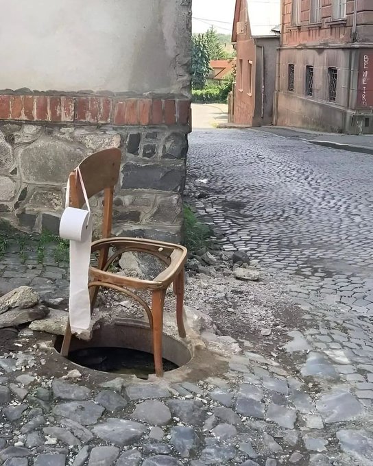 An old wooden chair sits over an open manhole on a cobblestone street, with a roll of toilet paper hanging from its backrest, with old buildings in the background.