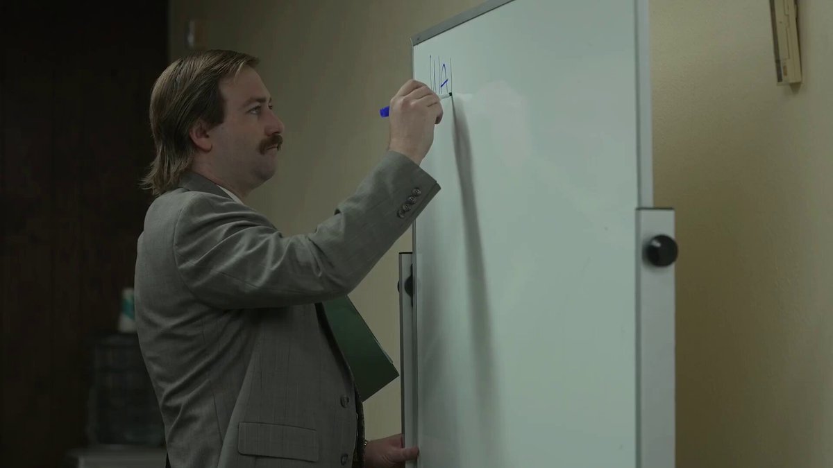 The image shows a man wearing a suit standing next to a whiteboard with blue writing on it. The man appears to be writing something, possibly a note or a message, as he holds a marker in his hand. The background of the image is blurred, but there are some indications that the setting might be an office environment.