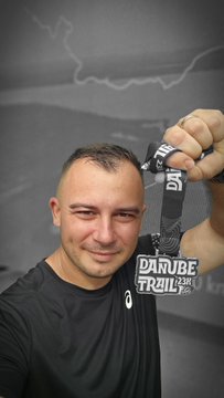 A smiling man wearing a black shirt holds up a 'DANUBE TRAIL' race medal.