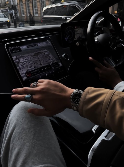 A close-up view of a car interior shows a person's hand, adorned with a silver watch and turquoise ring, resting near the center console while the dashboard displays "12:04" and a navigation map. The driver appears to be navigating urban streets, complementing the casual, on-the-go vibe of the "$SOL" crypto community's "gm" greeting.