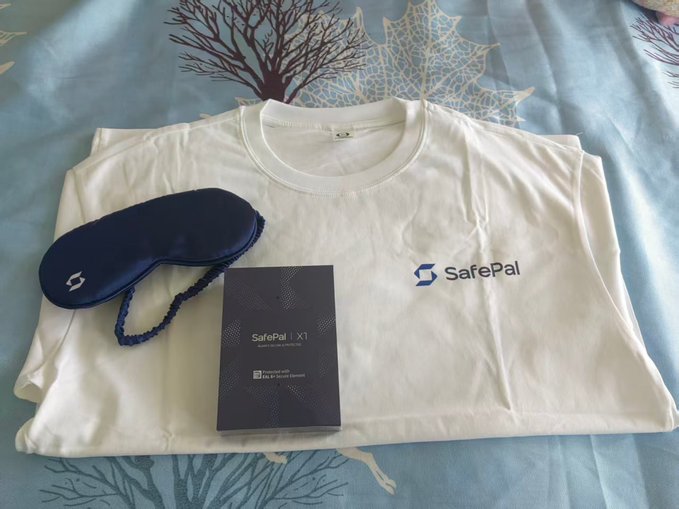 A folded white SafePal-branded t-shirt rests on a blue-patterned bedsheet, accompanied by a navy blue eye mask and a SafePal X1 hardware wallet box. The giveaway items pictured align with the post's promotion of 3 SafePal hardware wallets as part of the SosoValue co-branded giveaway.