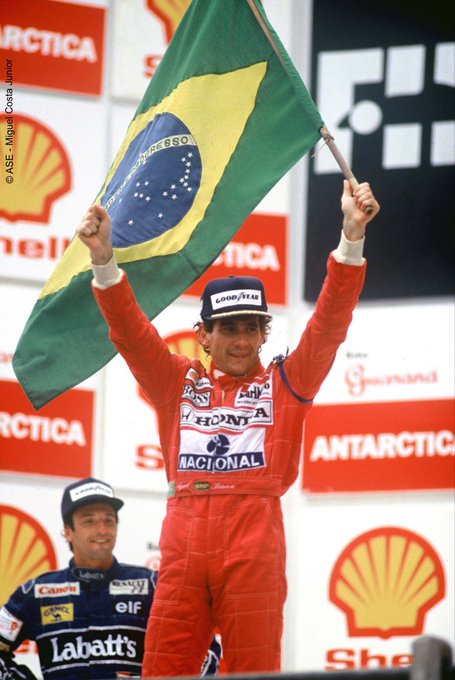 A Formula 1 driver in a red Honda/Nacional racing suit holds up a Brazilian flag on the podium, with another driver visible behind him and Shell and Antarctica sponsor logos in the background.