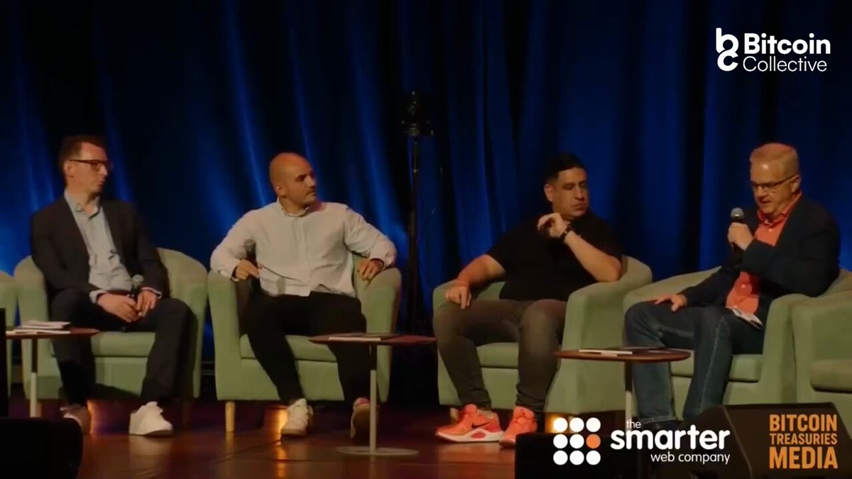 The image shows four men sitting around a table, engaged in conversation. They are all wearing suits and ties, suggesting that they might be attending an important event or discussing business matters. One of them has a microphone, indicating that he may be moderating the discussion or presenting information related to Bitcoin. The panelists appear to be well-dressed and focused on their conversation, which suggests that this is a formal gathering or meeting.