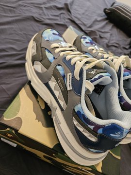 The image features a pair of blue and gray sneakers placed on top of a box, which appears to be from a brand called "Bape". The sneakers are positioned with their laces tied up neatly. In the background, there's a bed that is covered in a black blanket. There is also an item of clothing visible at the bottom right corner of the image.