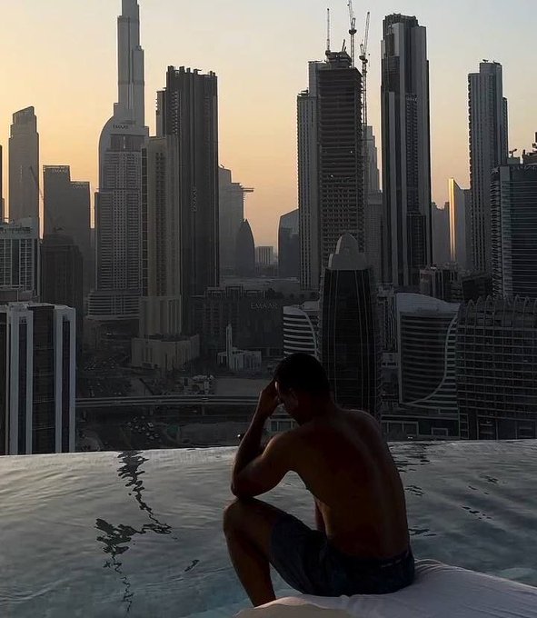 A shirtless man sits on the edge of an infinity pool overlooking a hazy city skyline of skyscrapers, including the Burj Khalifa, at sunset.