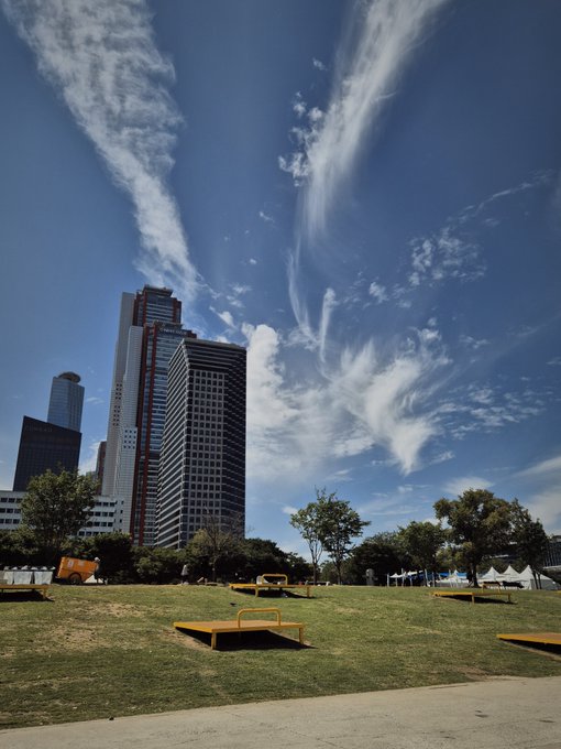 A park scene featuring a grassy area with wooden platforms in the foreground, with tall skyscrapers rising against a vivid blue sky streaked with wispy white clouds.