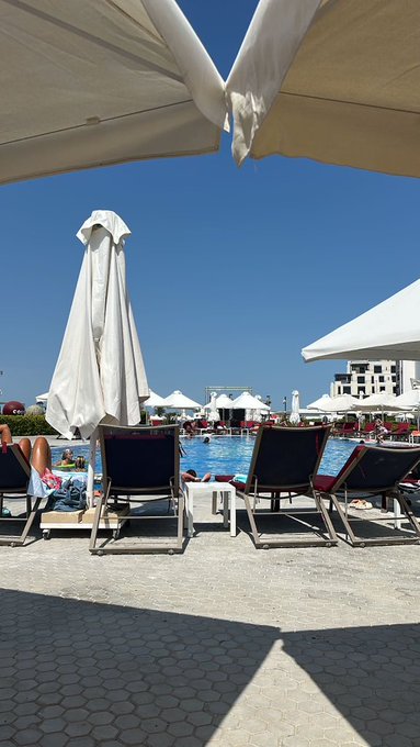 A sunny outdoor pool area with white sun umbrellas, lounge chairs, and people swimming in a pool under a clear blue sky.