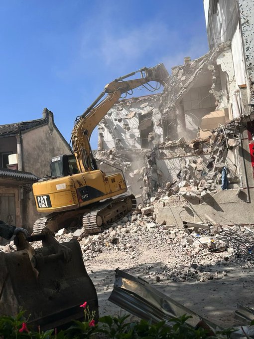 A yellow CAT 312C excavator demolishes a concrete building, with rubble and dust scattered around the work site.
