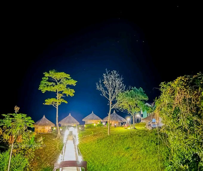 A night scene showing thatched-roof huts illuminated by lights, with trees in the foreground and a wooden bridge leading towards the buildings under a dark sky.