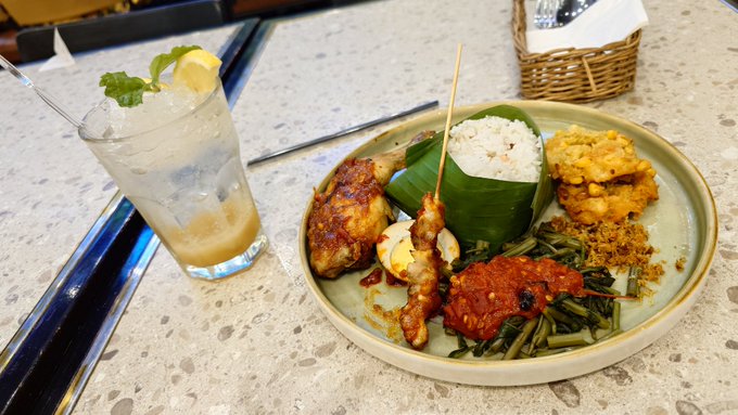 A photo of an Indonesian-style meal plate featuring chicken satay skewers, rice wrapped in banana leaf, sambal, vegetables, and fritters, accompanied by a glass of iced drink with lemon and mint.