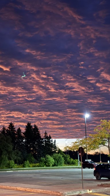A dramatic sunset or sunrise sky with pink and orange clouds illuminating a parking lot area, with trees silhouetted against the colorful sky and a streetlight glowing.