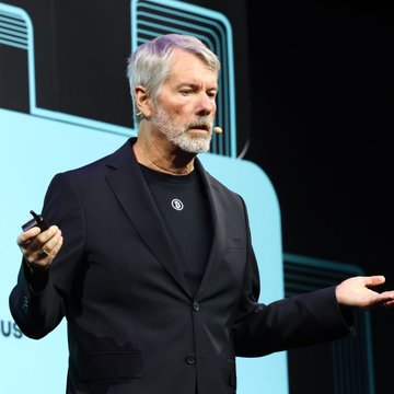 A gray-bearded man in a black blazer and t-shirt speaks on stage with a headset microphone, gesturing with both hands.