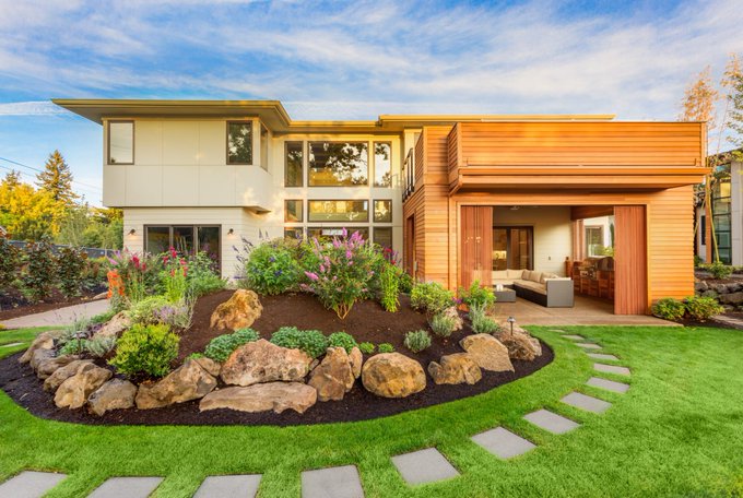 A modern two-story home features mixed exterior materials of wood paneling, stucco, and large windows, with a landscaped rock garden, stepping stone path, and open patio in the foreground.