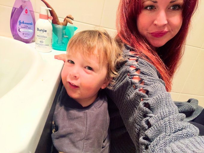 A selfie of a woman with red hair and a young child sitting next to a bathtub with Johnsons and Bunjie baby products visible on the ledge.