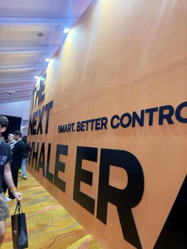 A view of a yellow exhibition or hallway wall displaying the text 'NEXT SCALE ER' and 'SMART, BETTER CONTRO...' with people walking past.